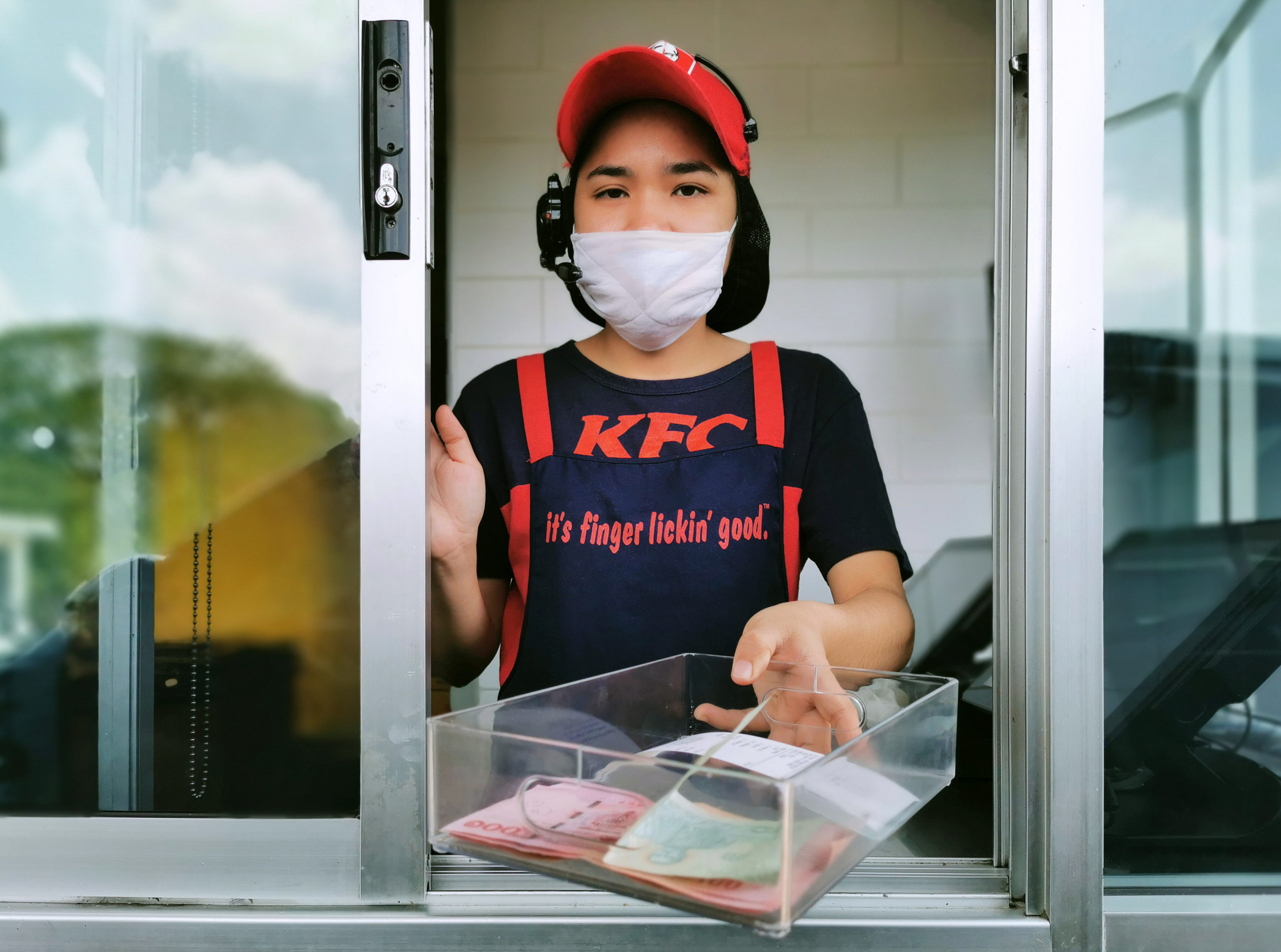 KFC fast food cashier in drive thru service waring hygiene face mask to protect coronavirus pandemic or covid-19 virus outbreak is giving money change to customer.